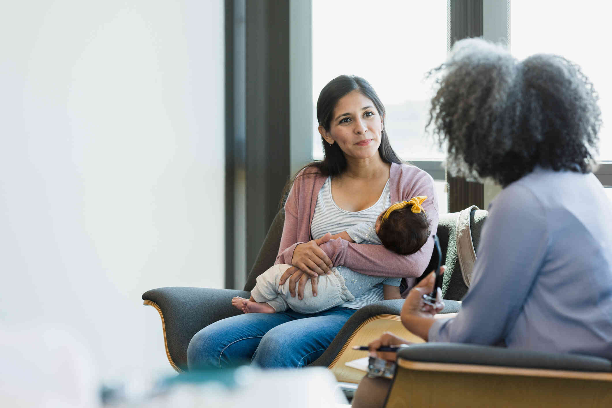 A woman sits in a chair and holds her infant in her arms as she talks to the female therapist sitting across from her.